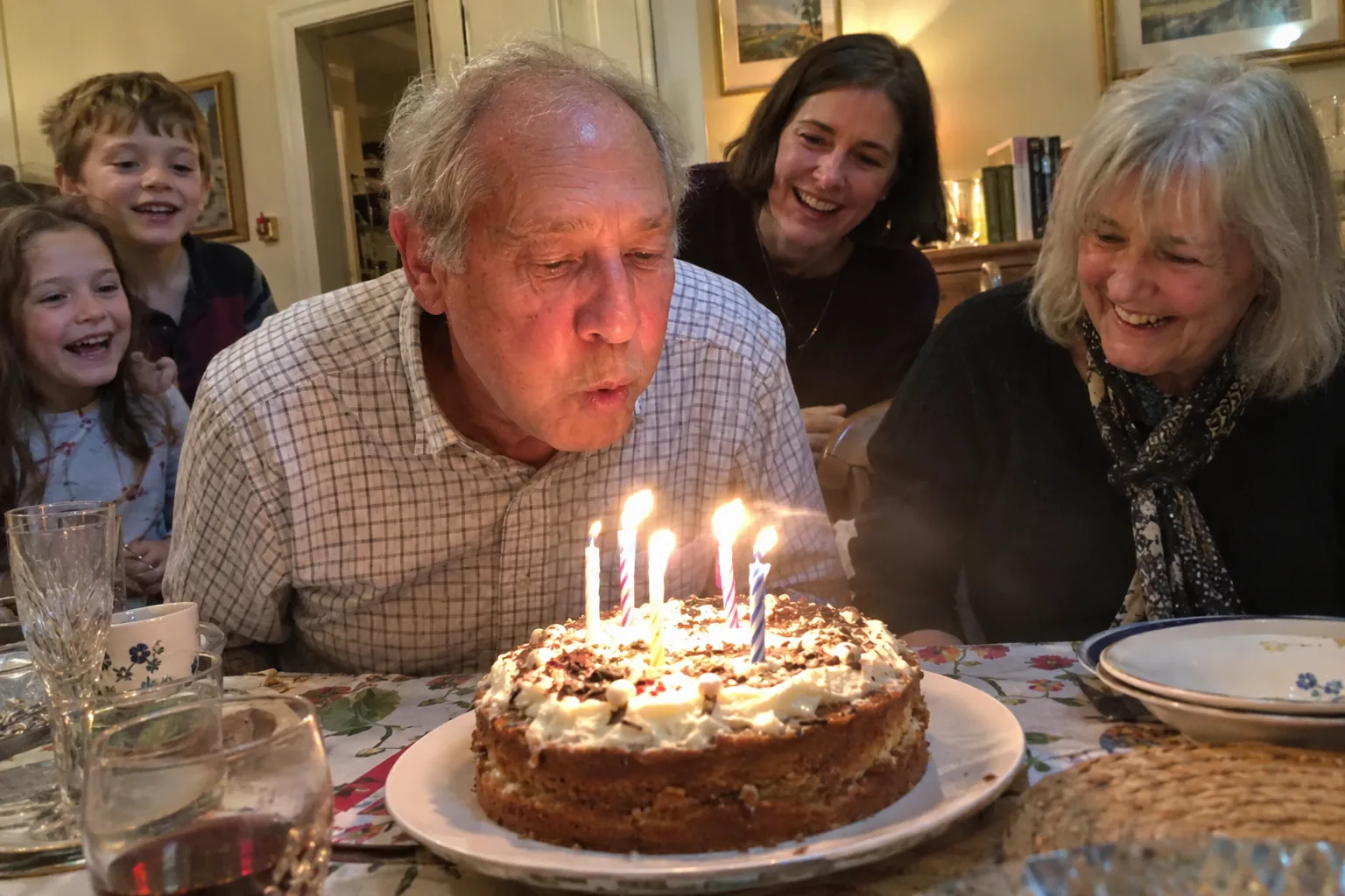 An older man blowing out birthday candles while relatives smile behind him.
