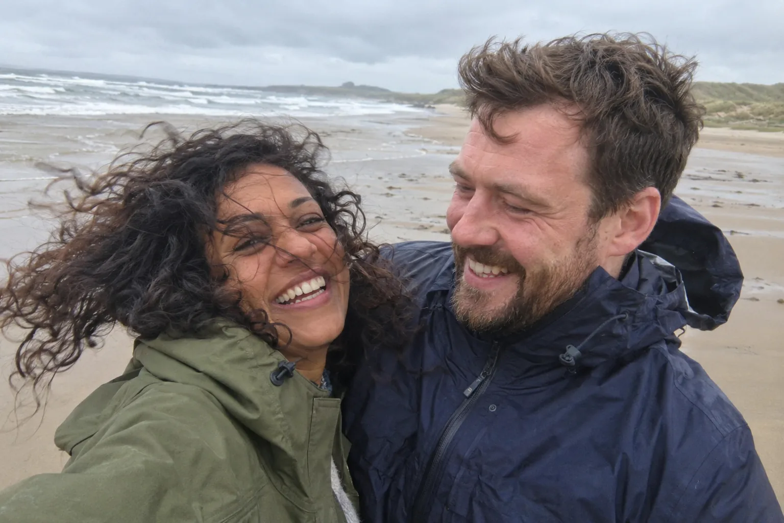 A smiling couple in jackets on a windswept Northumberland beach.